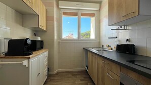 A compact kitchen with wooden cabinets, a microwave, a toaster, and a window with a view of the sky. - RIVÉA - 2-bedroom apartment near the ocean (Mimizan)