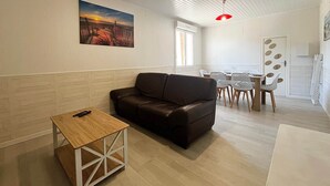 A living room with a brown sofa, a wooden coffee table, a dining area with a white table and chairs, and a painting on the wall. - RIVÉA - 2-bedroom apartment near the ocean (Mimizan)