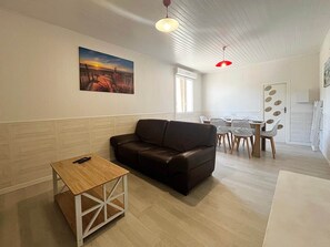 Ein Wohnzimmer mit braunem Sofa, einem Holztisch, einem Essbereich mit weißem Tisch und Stühlen sowie einem Bild an der Wand.