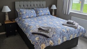 Une chambre à coucher avec une tête de lit capitonnée, une literie à motifs floraux, deux lampes de chevet et une vue sur l’extérieur.