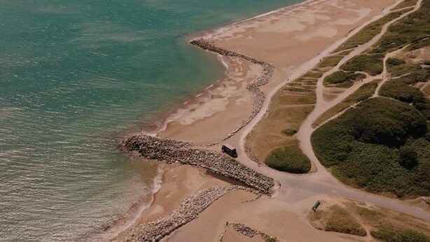 Vue aérienne d’une zone côtière comprenant une plage, un mur de pierres et une route menant à un espace verdoyant.