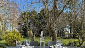 An outdoor dining table set with plates, cutlery, and glasses, surrounded by trees and greenery. - Gîte in Baie de Somme – L'Ecurie (Cayeux-sur-Mer)