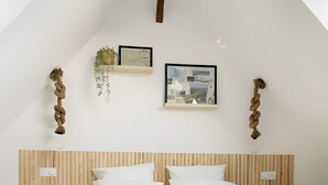A modern bedroom with a sloped ceiling, a wooden headboard, a large bed with white bedding, two bedside tables, and wall-mounted decor including a framed map and a plant. - Gîte in Baie de Somme – L'Ecurie (Cayeux-sur-Mer)