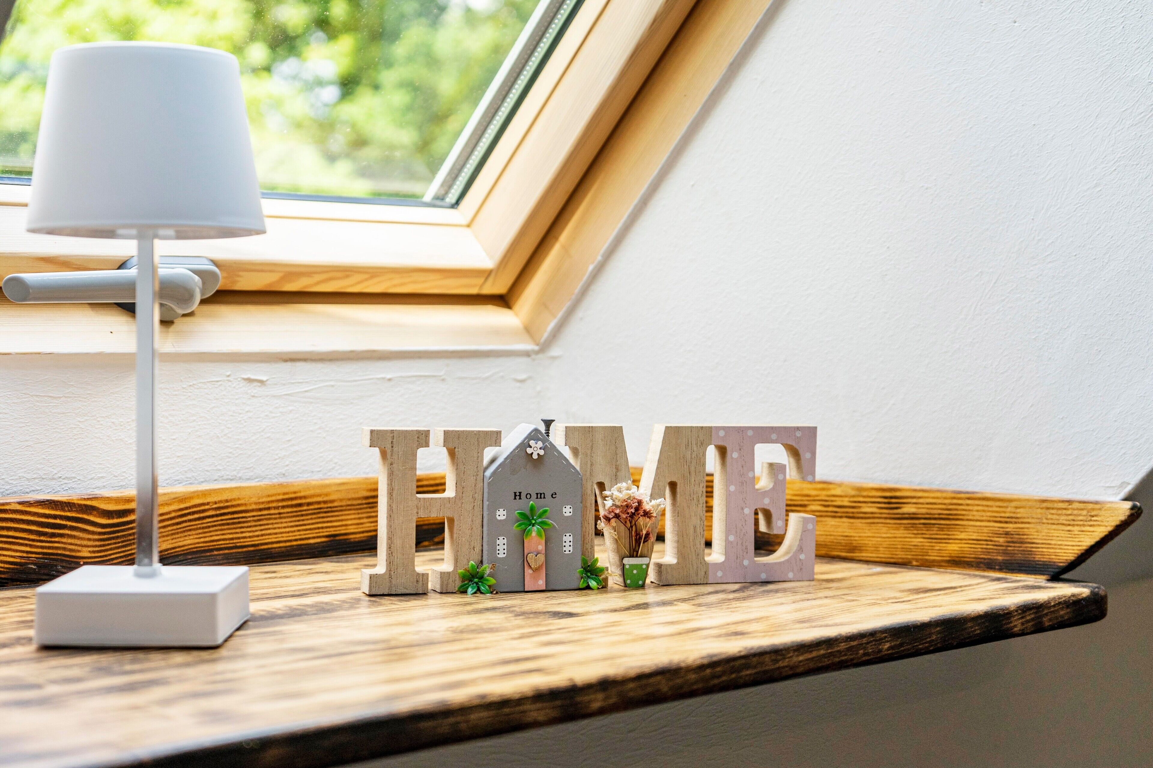 A window sill with a lamp, a wooden shelf, and decorative letters spelling 'HOME' with a small house model and plants.
