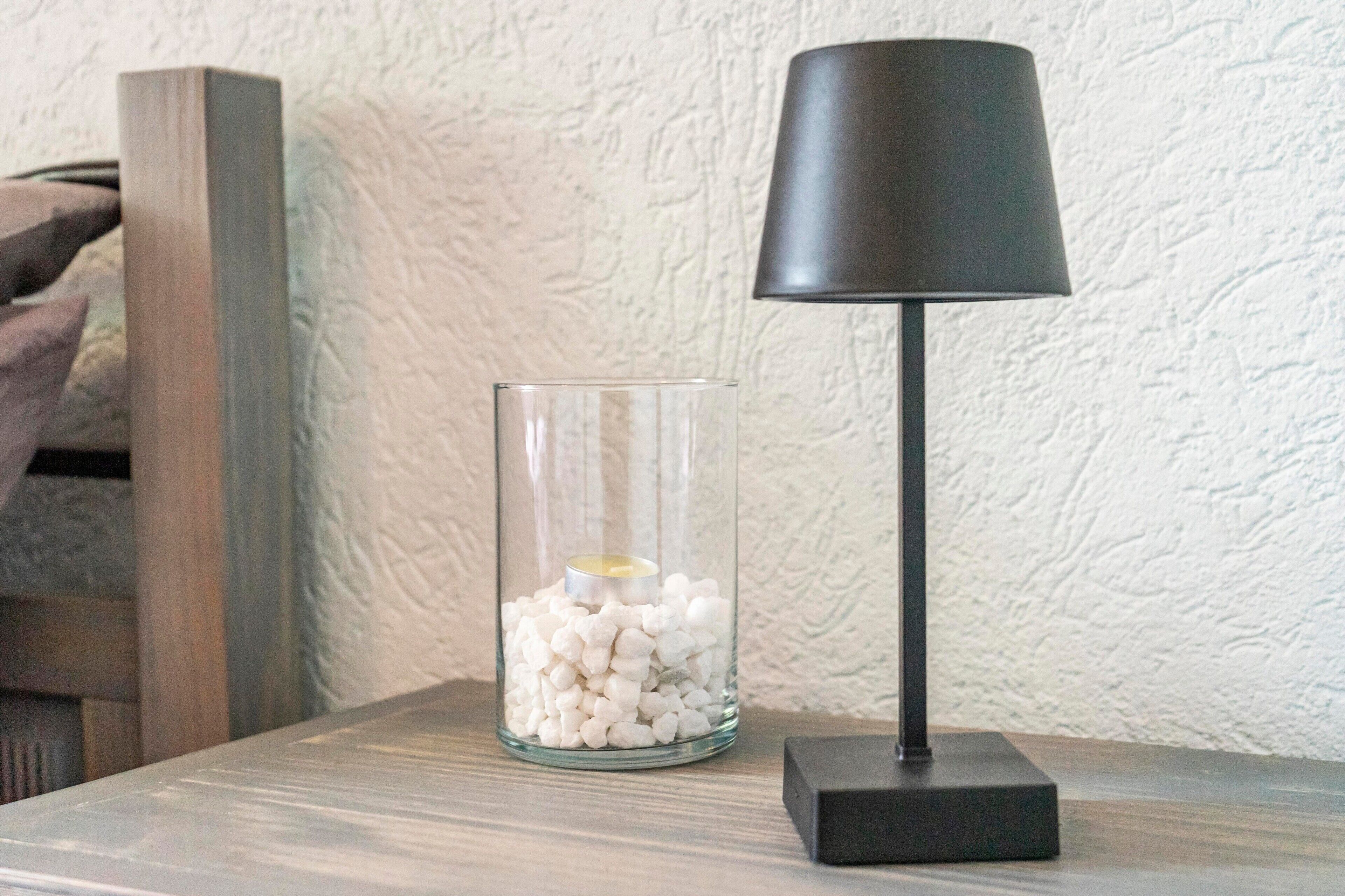 Un photophore en verre contenant des petits galets blancs et une bougie, placé sur une surface en bois à côté d’une lampe noire à la base carrée.