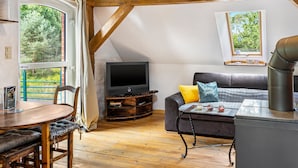 Una sala de estar acogedora con techo de madera, un televisor sobre un mueble de madera, un sofá con cojines, una mesa de comedor con sillas y vistas a zonas verdes.