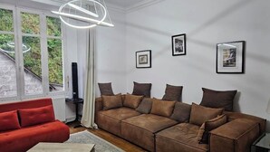 A living room with a sectional sofa, a coffee table, and a large window with curtains. - 90-square-meter apartment in the city center, 10 minutes from the train station, 200 meters from the cathedral (Quimper)
