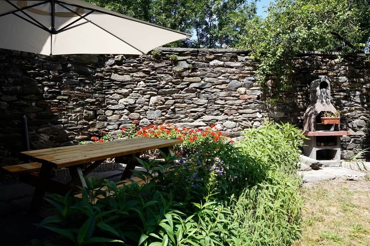 A stone wall, a wooden picnic table, and a barbecue grill in a garden setting.