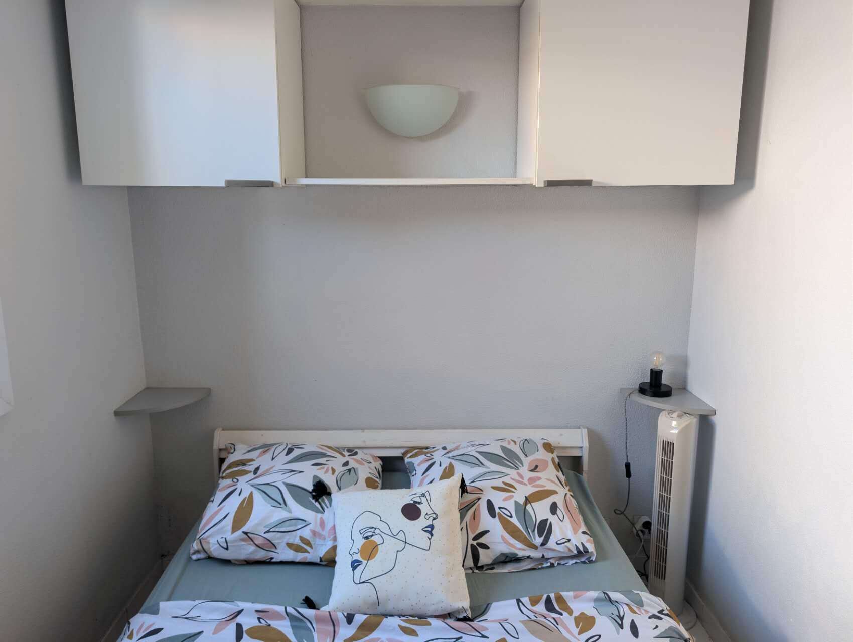 Ein kleines, ordentlich eingerichtetes Schlafzimmer mit einem Einzelbett, einem Nachttisch, einem Wandregal und einer Wandleuchte.
