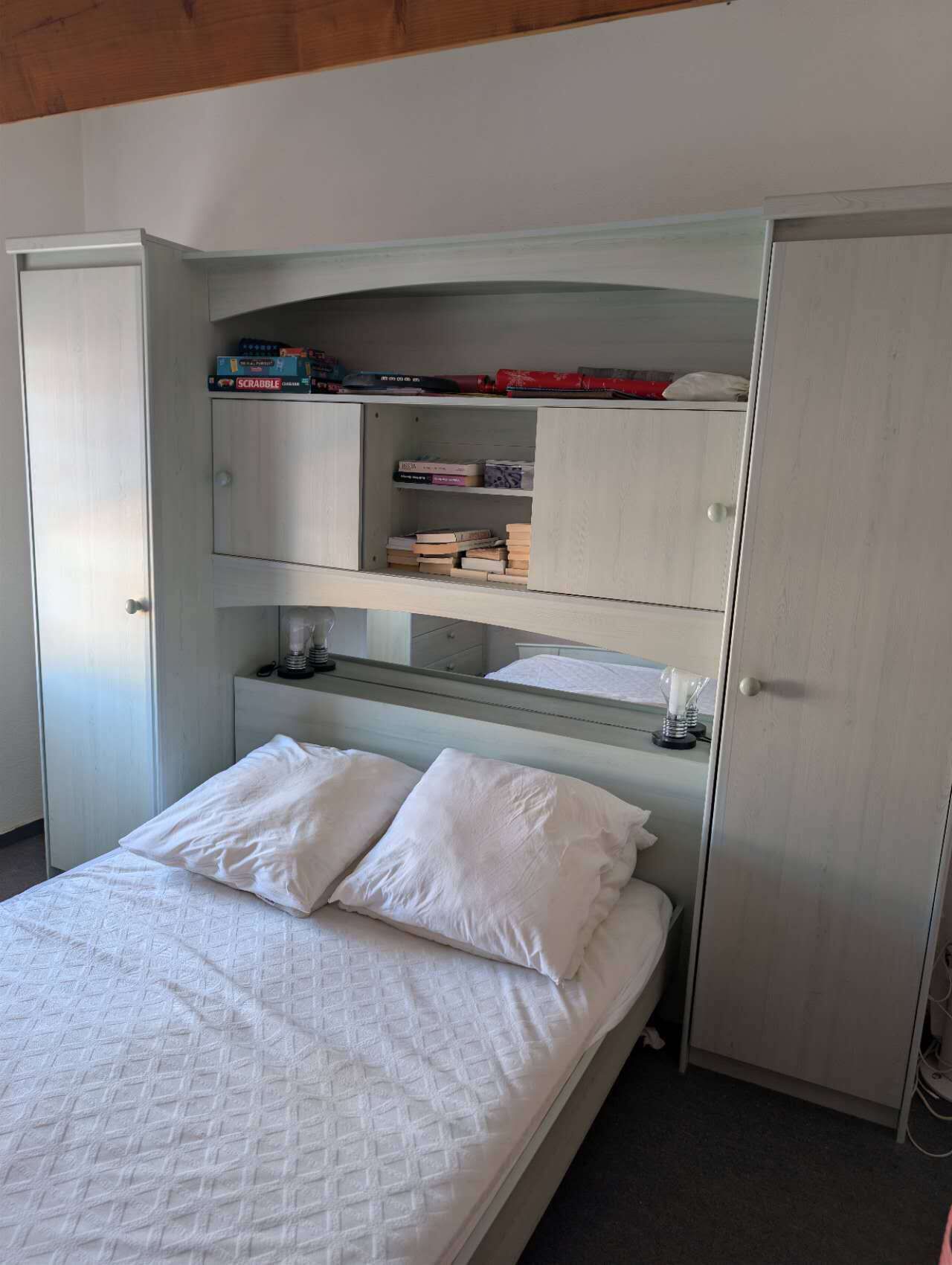 A bed with white bedding, a bookshelf with books, and a wardrobe with a mirror.