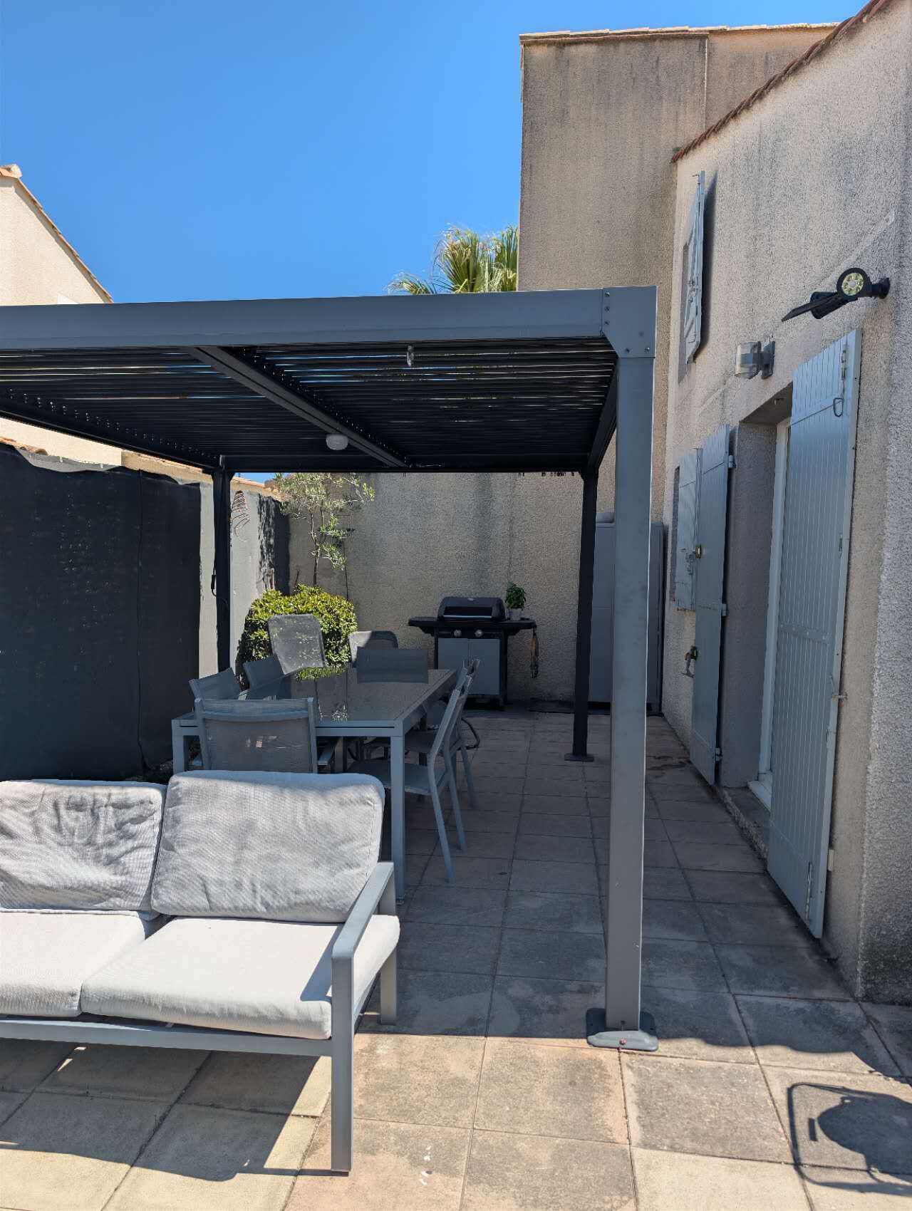 Eine überdachte Terrasse im Freien mit Tisch, Stühlen, einem Grill und einer Couch.