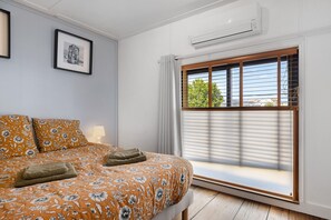 Un dormitorio con cama, ventana con persianas, unidad de aire acondicionado y un cuadro en la pared.