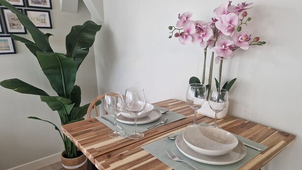 Un tavolo da pranzo apparecchiato con piatti, bicchieri e posate, arricchito da una pianta in vaso e fiori di orchidea.