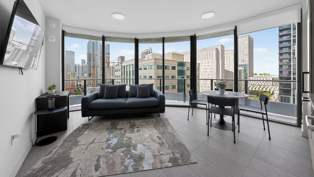 Una sala de estar moderna con sofá, una mesa de centro y un comedor, con grandes ventanales y vista a la ciudad.