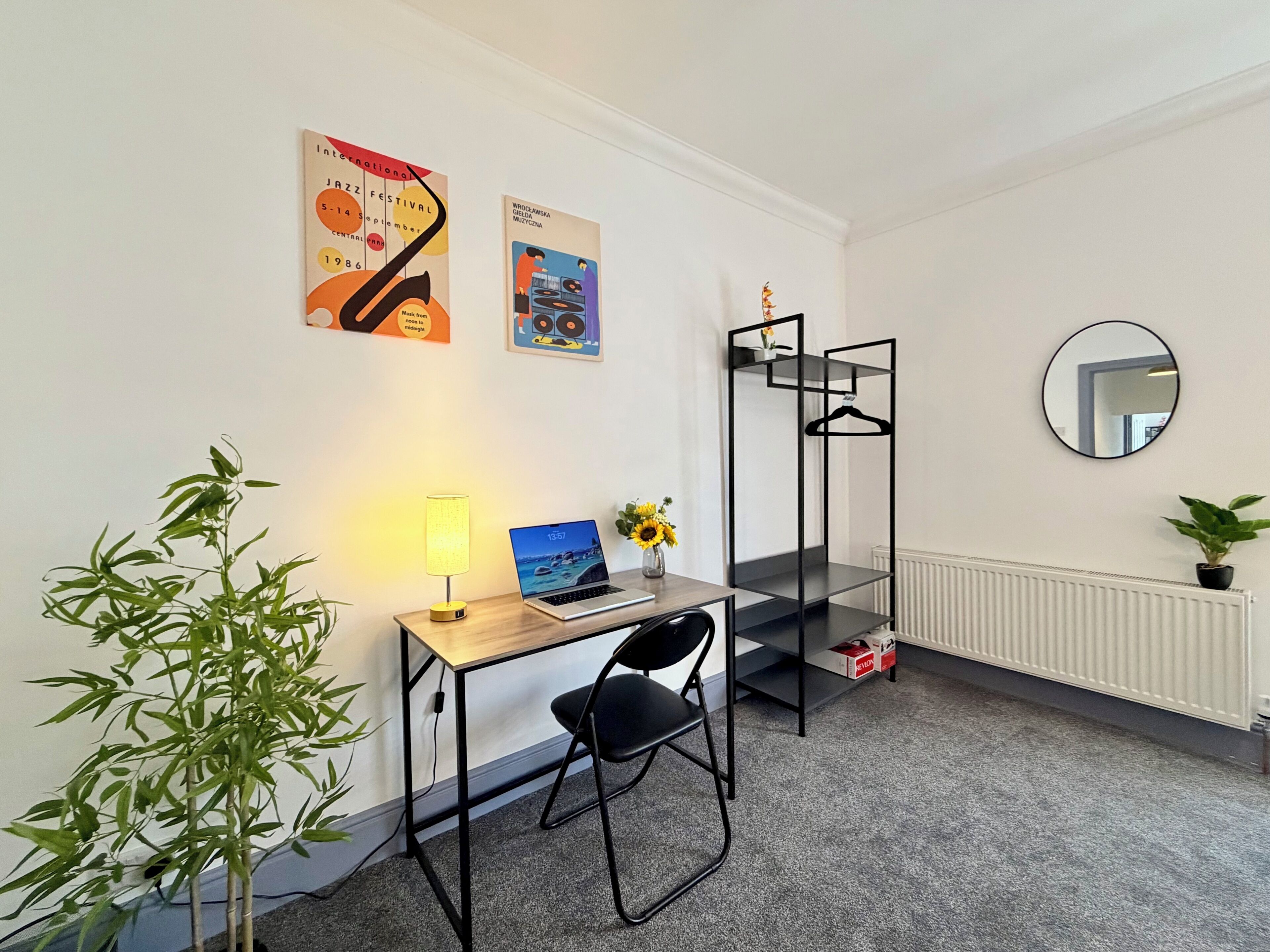 A room with a desk, chair, lamp, laptop, two posters, a mirror, a radiator, and a plant.