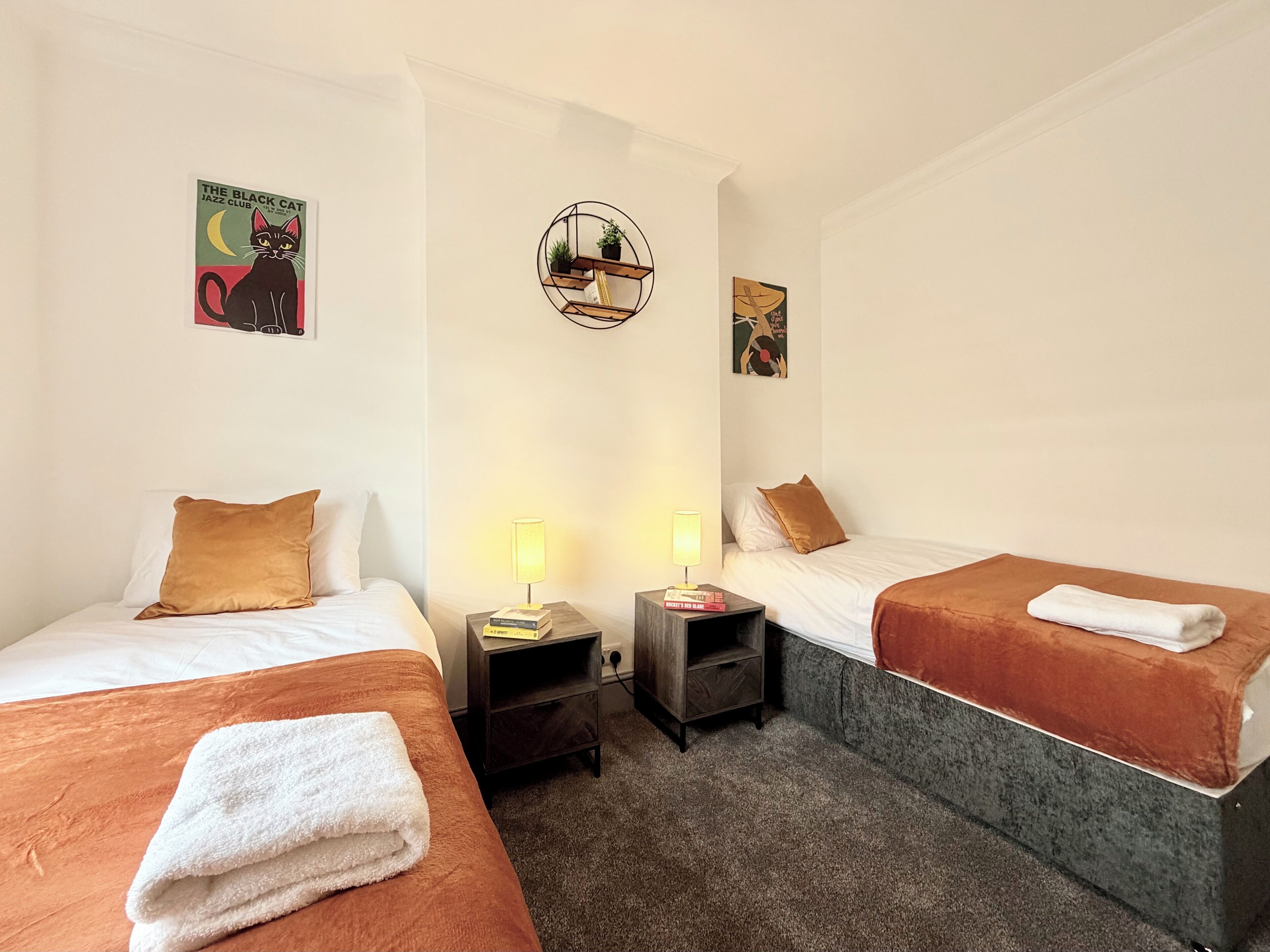 A hotel room with two beds, a nightstand with a lamp, a small shelf with books, and a wall clock.