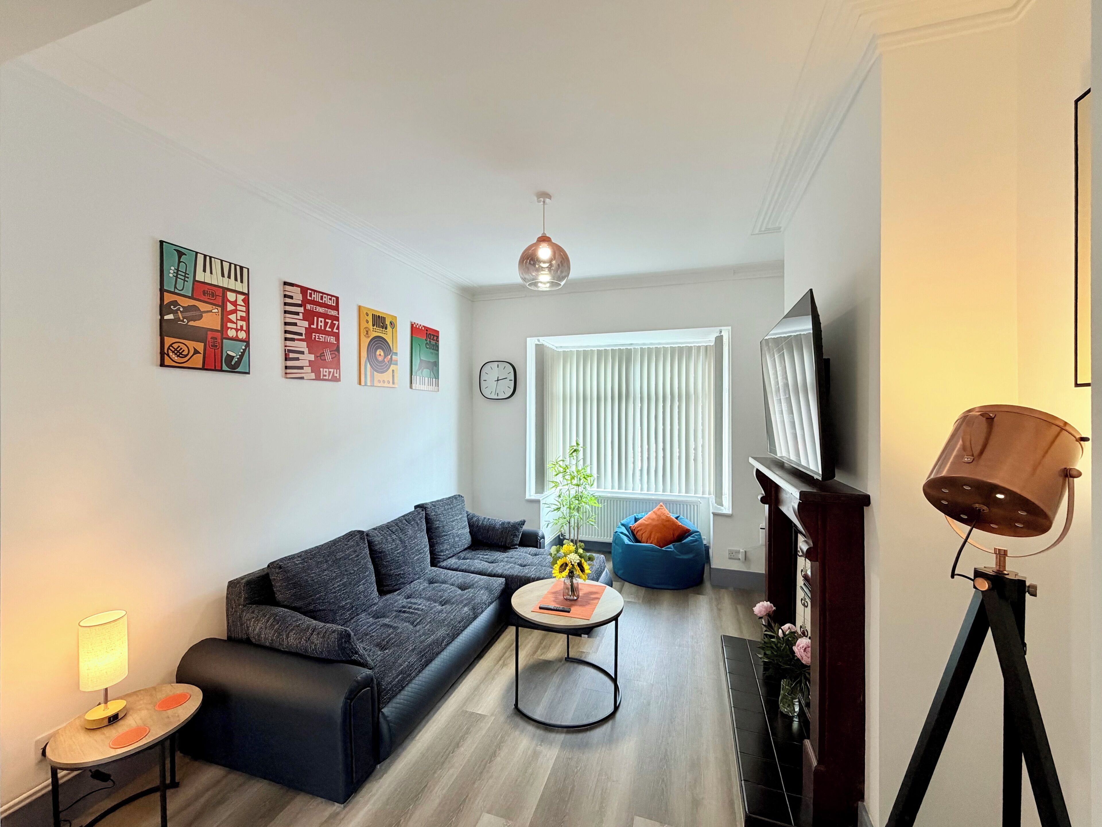 A modern living room with a sectional sofa, a small round table with a lamp, a blue bean bag chair, a potted plant, and a tripod lamp.