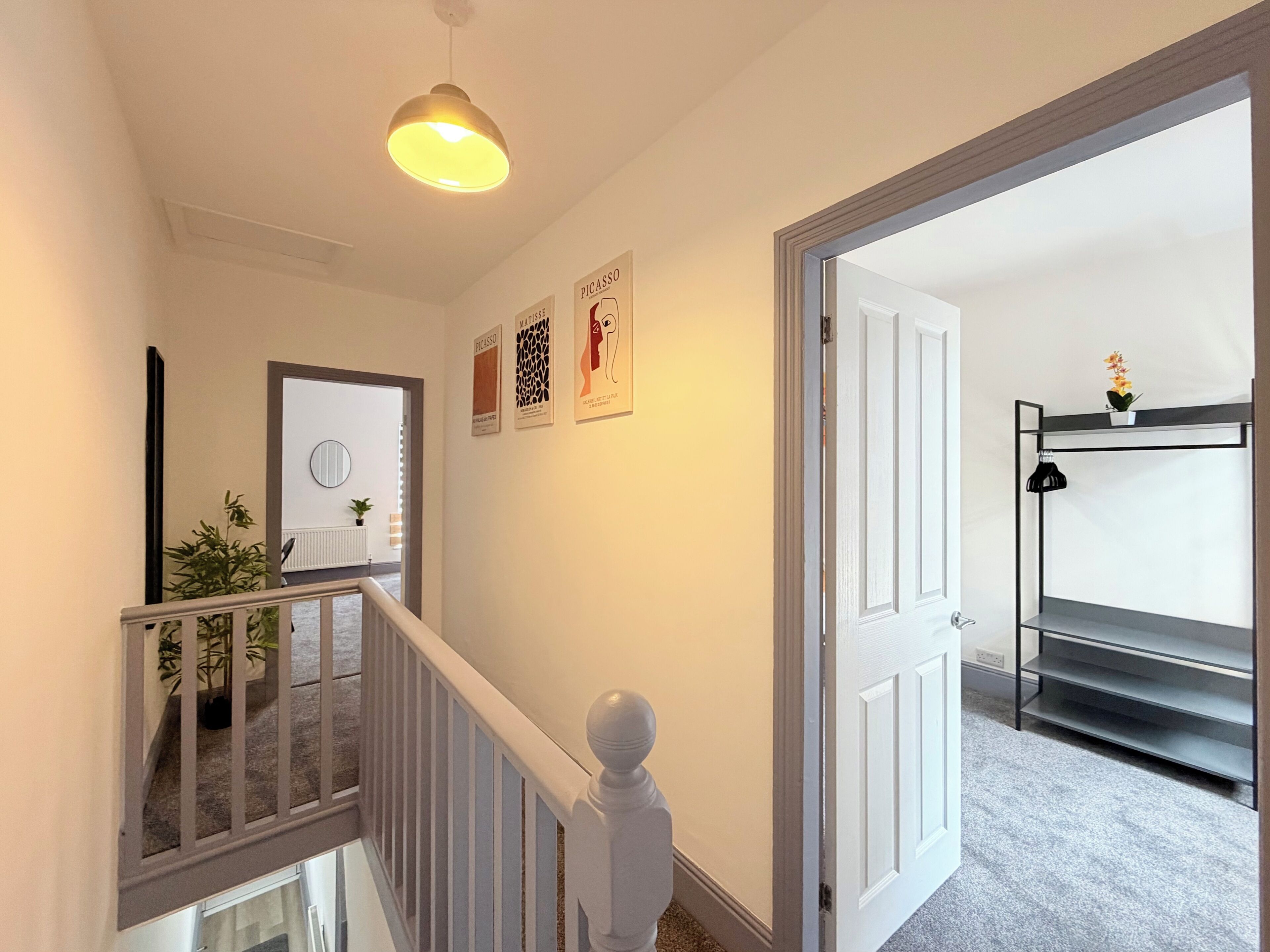 A hallway with a staircase, a hanging light fixture, a door leading to another room, and wall-mounted artwork.