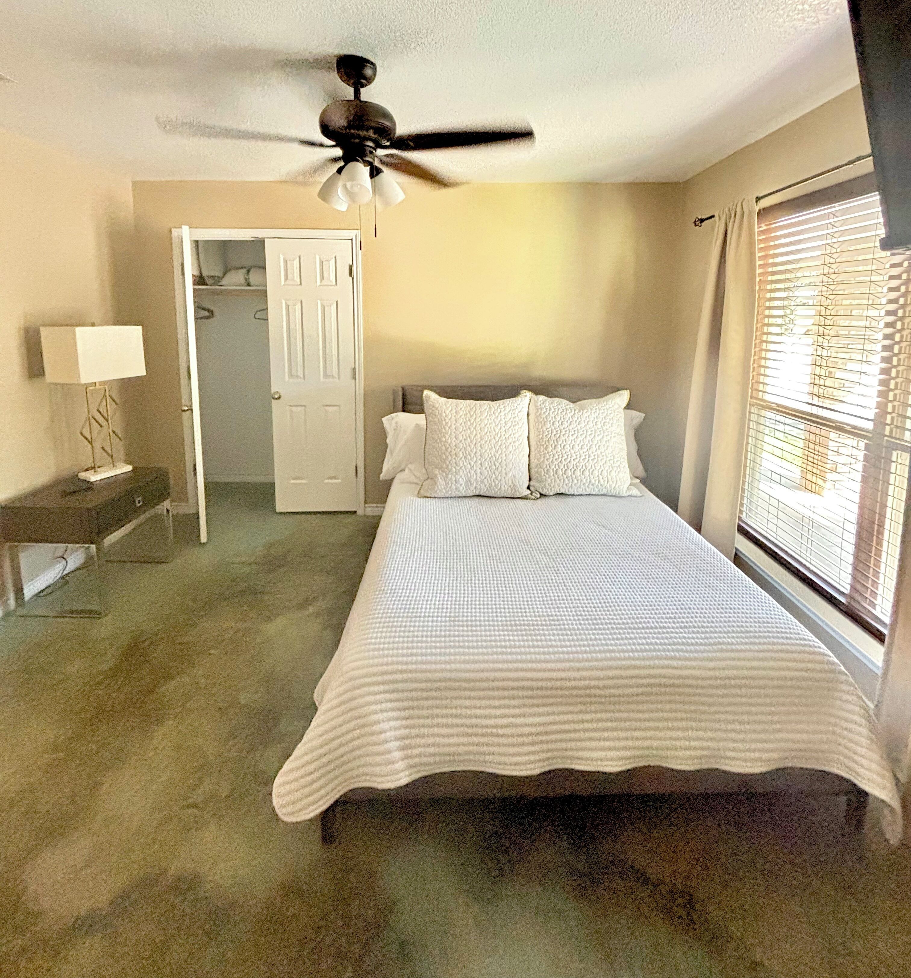 A bedroom with a bed, a ceiling fan, a nightstand, a lamp, a door, and a window with blinds.