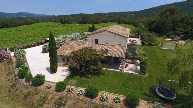 Une maison avec un toit en tuiles, entourée d’une pelouse et d’un jardin bien entretenus, et on aperçoit un court de tennis au loin.