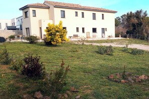 A two-story building with a flat roof, multiple windows, and a well-maintained lawn with small shrubs and a yellow flowering tree. - Apartment (Marseillan)