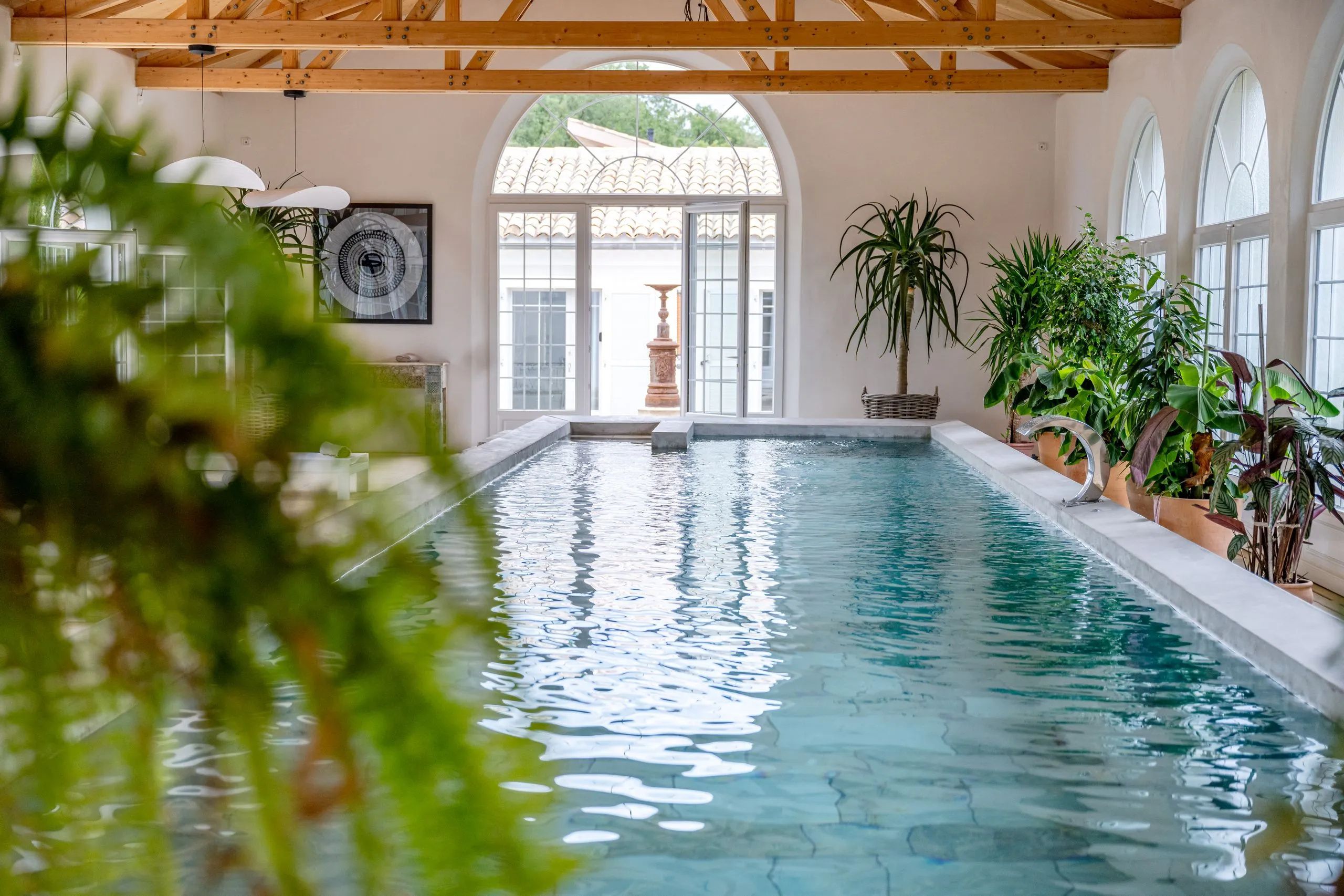 Een binnenzwembad met helder water, houten balken in het plafond en potplanten rondom het zwembad.