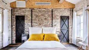 Une chambre avec un mur en briques, un lit avec des coussins blancs et jaunes, un plancher en bois et une fenêtre avec des rideaux.