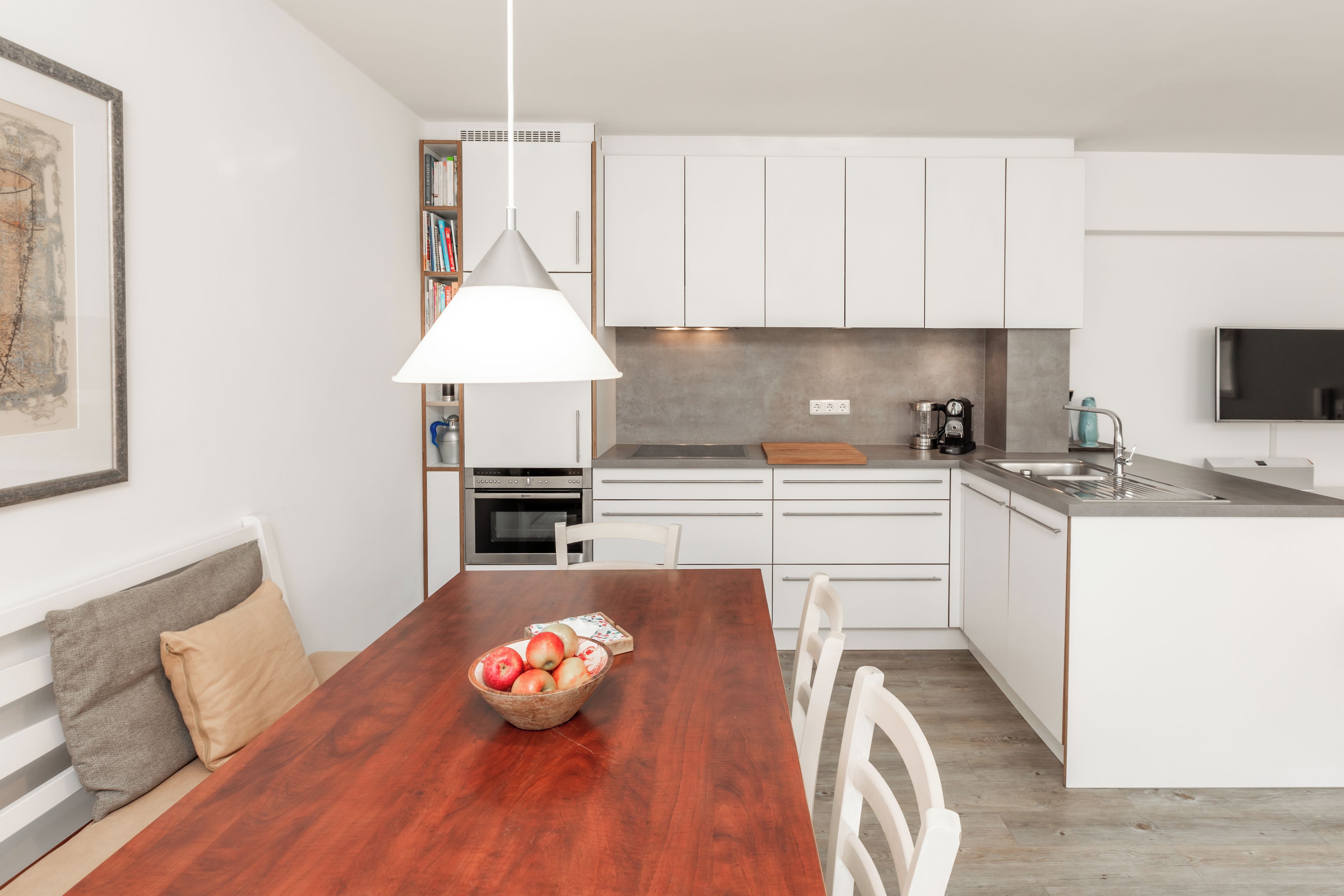 Une cuisine moderne avec des meubles blancs, une table à manger en bois sur laquelle se trouve un saladier rempli de fruits, et un luminaire suspendu.