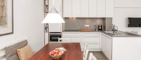 Une cuisine moderne avec des meubles blancs, une table à manger en bois sur laquelle se trouve un saladier rempli de fruits, et un luminaire suspendu.