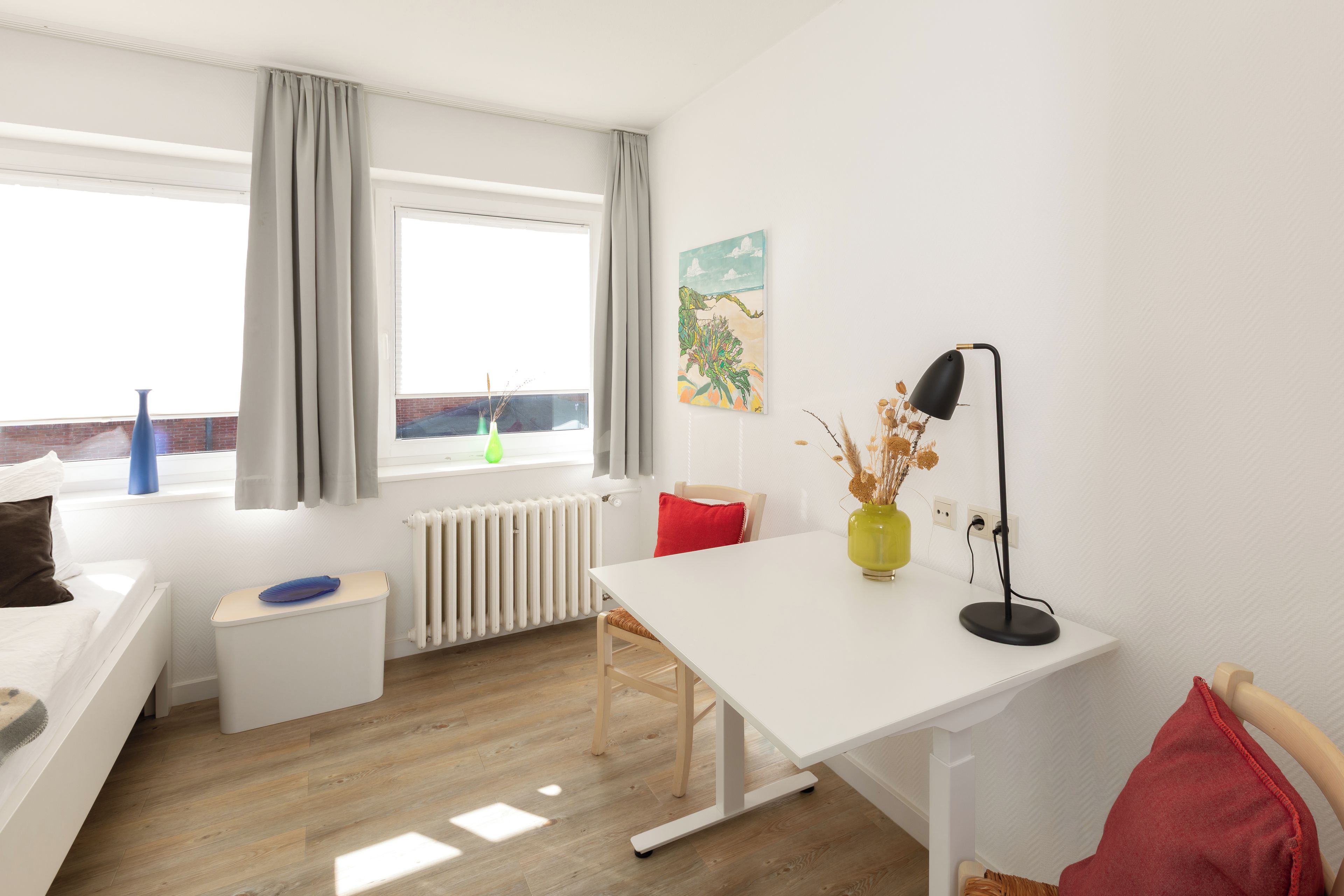Een moderne, goed verlichte kamer met een wit bureau, een stoel, een radiator, een raam met gordijnen en een schilderij aan de muur.