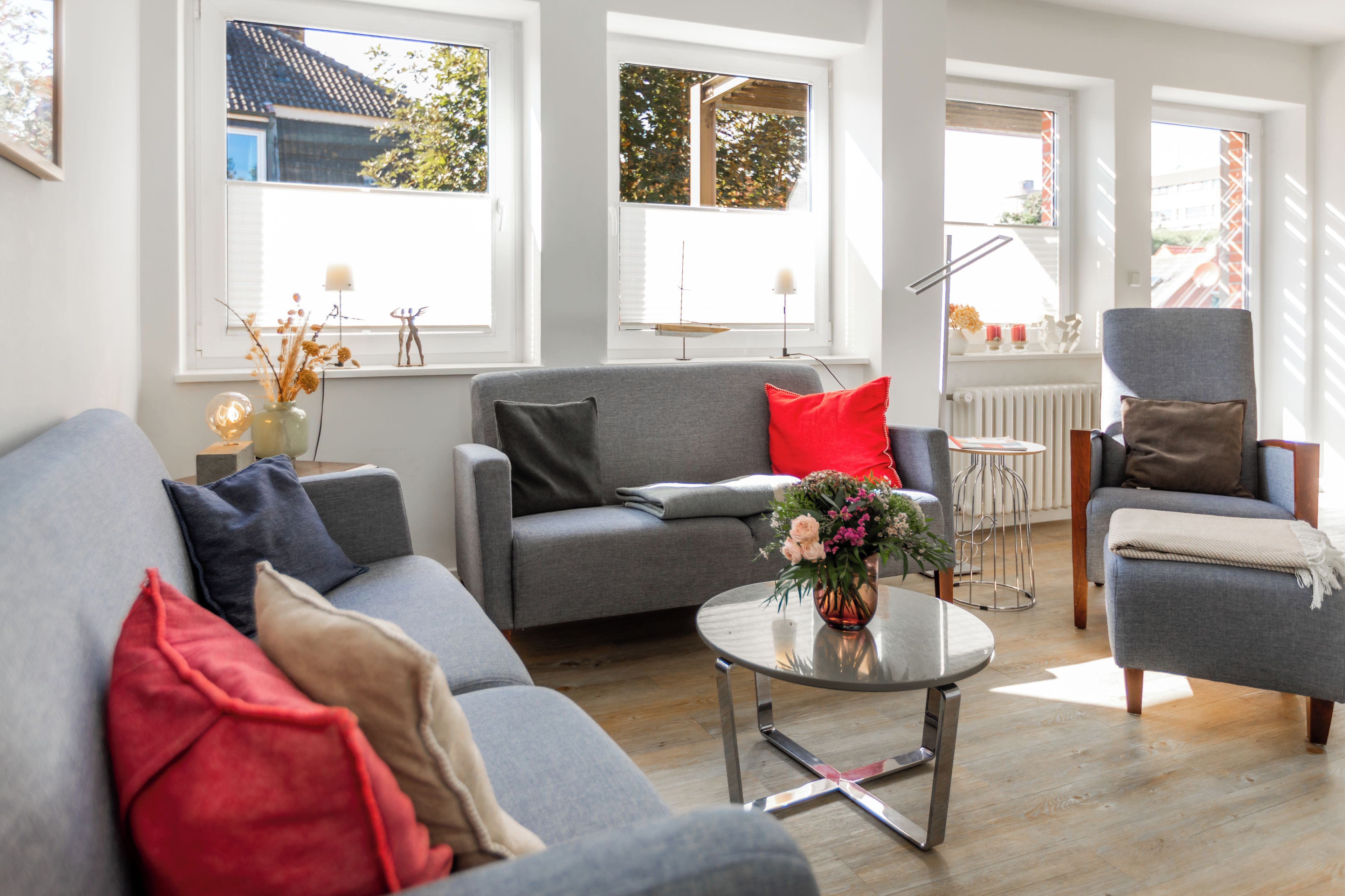 A modern living room with a grey sofa, a red pillow, a small round table with a vase of flowers, and a grey armchair.