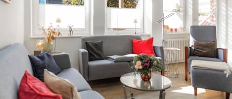 A modern living room with a grey sofa, a red pillow, a small round table with a vase of flowers, and a grey armchair.