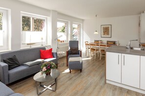 Un espacio de estar moderno con un sofá gris, una pequeña mesa redonda con un florero, una zona de comedor con una mesa y sillas de madera, y una cocina con armarios blancos y un lavamanos.