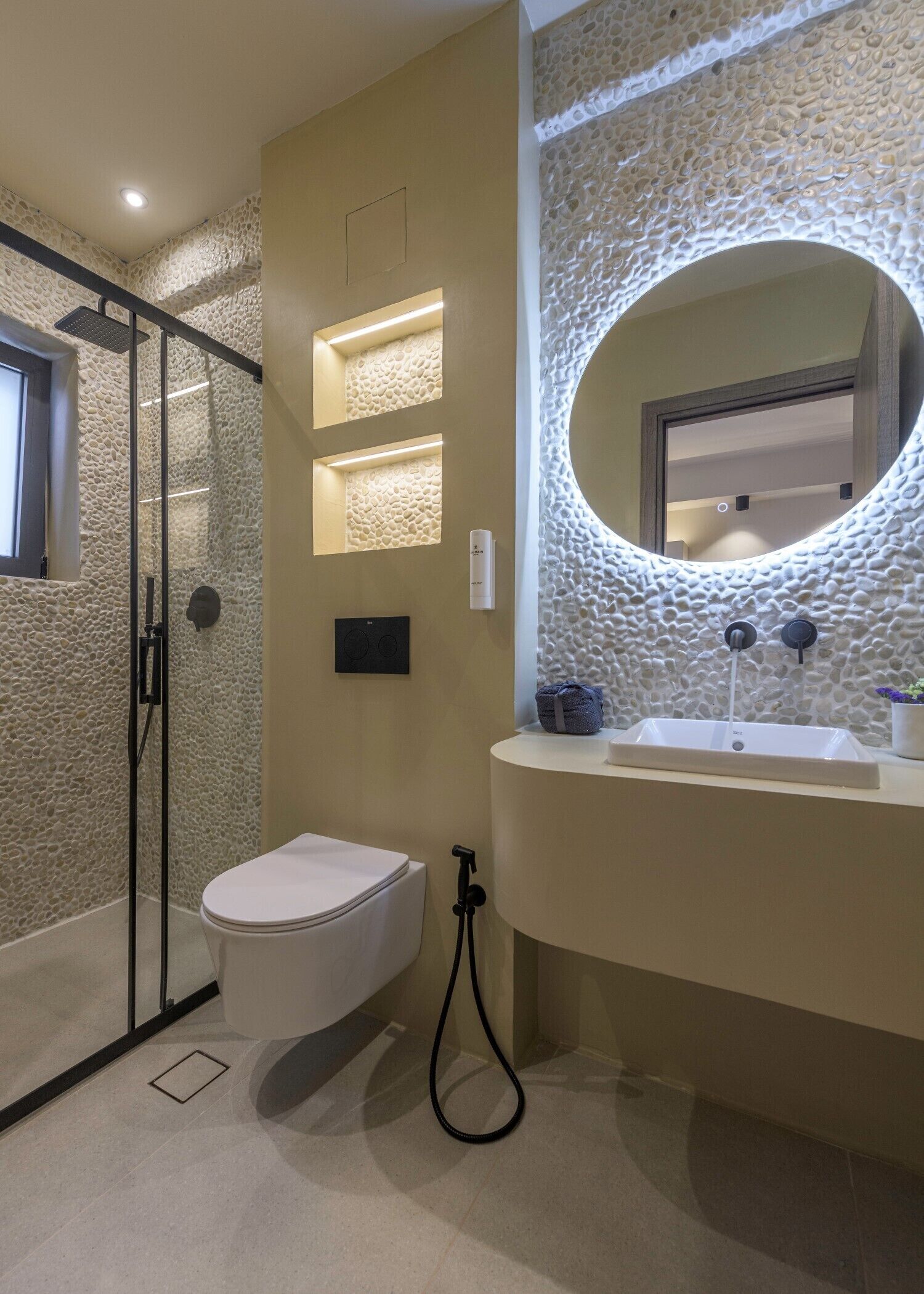Une salle de bain avec un mur à l’aspect pierre, une douche en verre, des toilettes, un lavabo avec un miroir et un éclairage encastré.