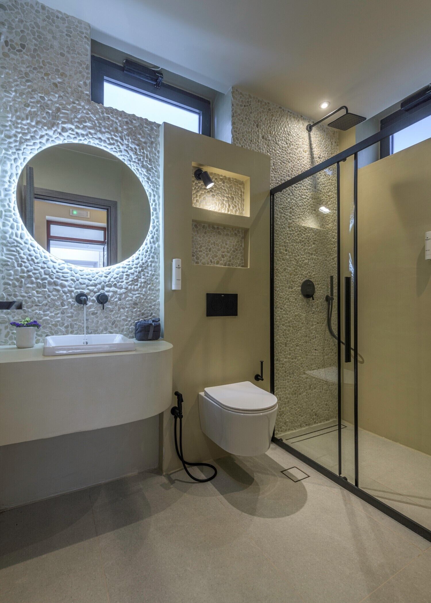Un baño moderno con pared de textura de piedra, espejo redondo, lavamanos blanco, inodoro y zona de ducha con mampara de vidrio.