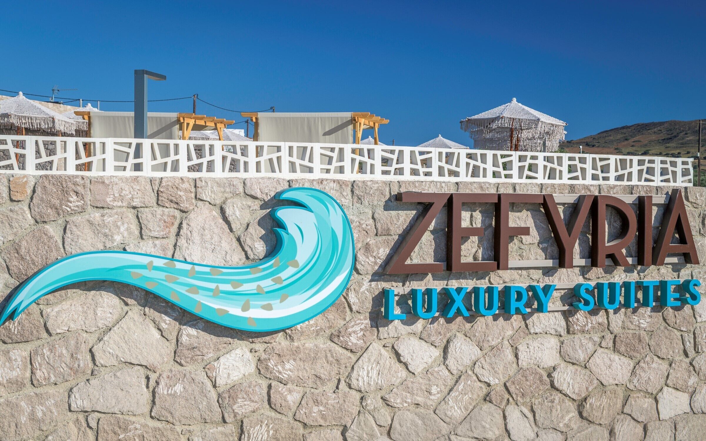 Une enseigne pour les Zefyria Luxury Suites, avec un motif stylisé en forme de vague et un mur en pierre en arrière-plan.