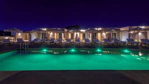 Un área de piscina nocturna con sillones de descanso y sombrillas, rodeada de muros de piedra y un edificio de piedra.