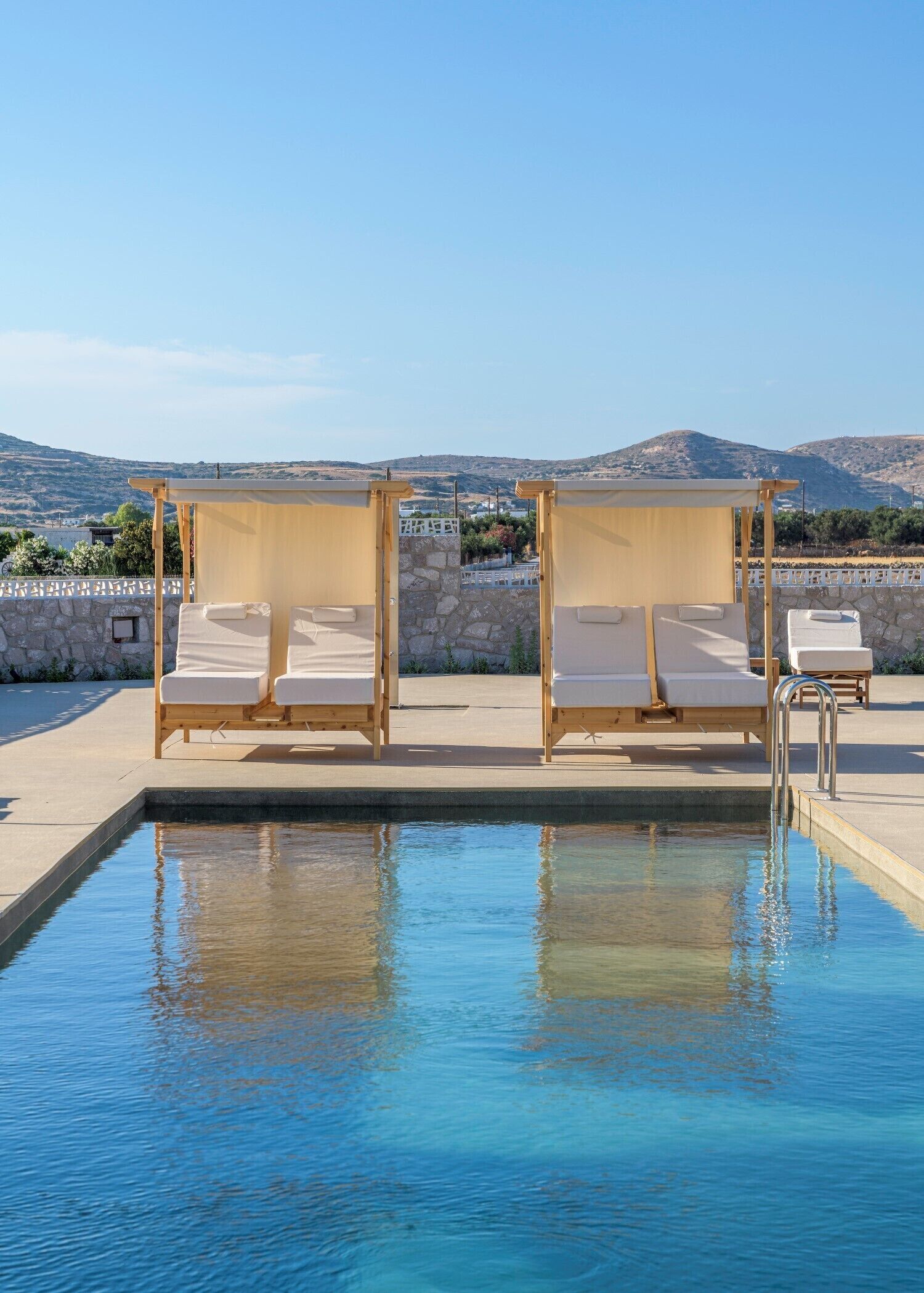 A pool with two sun loungers, a stone wall, and a view of hills.