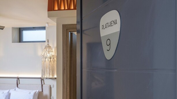 A modern hotel room with a bed, bedside table, and a door labeled 'PLATHIEN 9'.