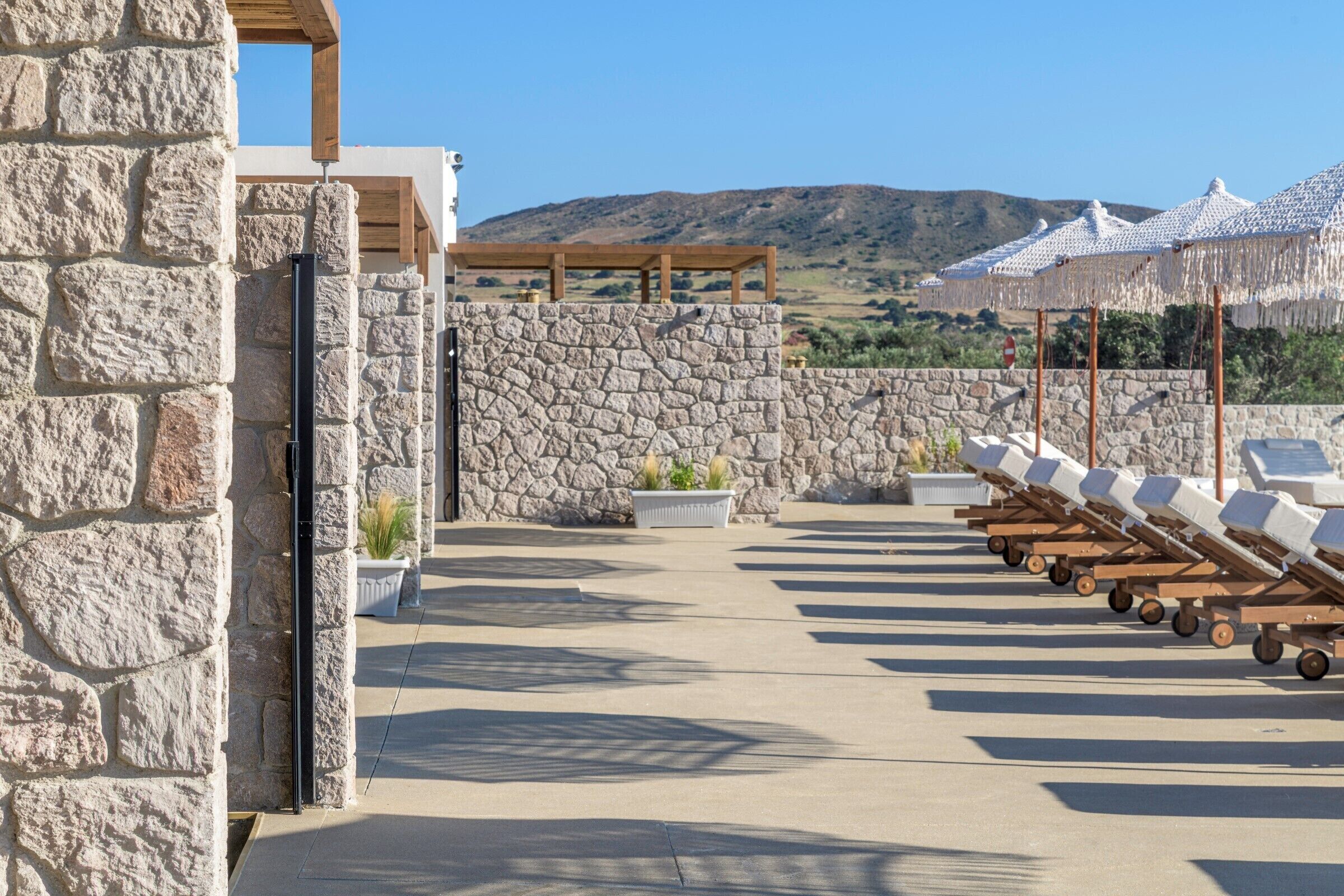 Un edificio de piedra con una pérgola de madera, mesas al aire libre con sombrillas y vistas a las colinas.