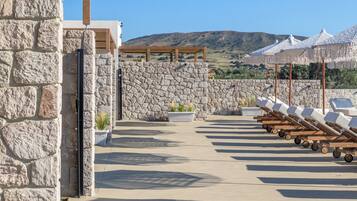 Un edificio de piedra con una pérgola de madera, mesas al aire libre con sombrillas y vistas a las colinas.