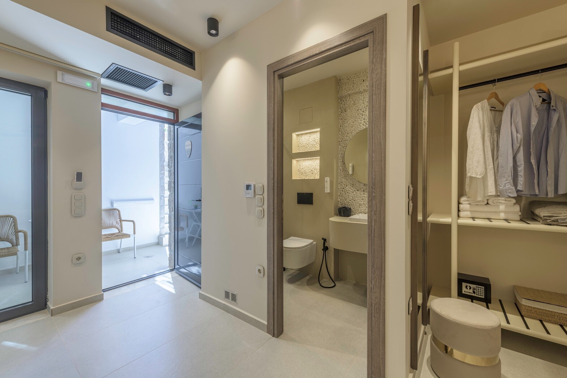 Ein modernes Hotelzimmer mit Glastür zum Balkon, einem Badezimmer mit Toilette und Waschbecken sowie einem begehbaren Kleiderschrank mit Kleidung.