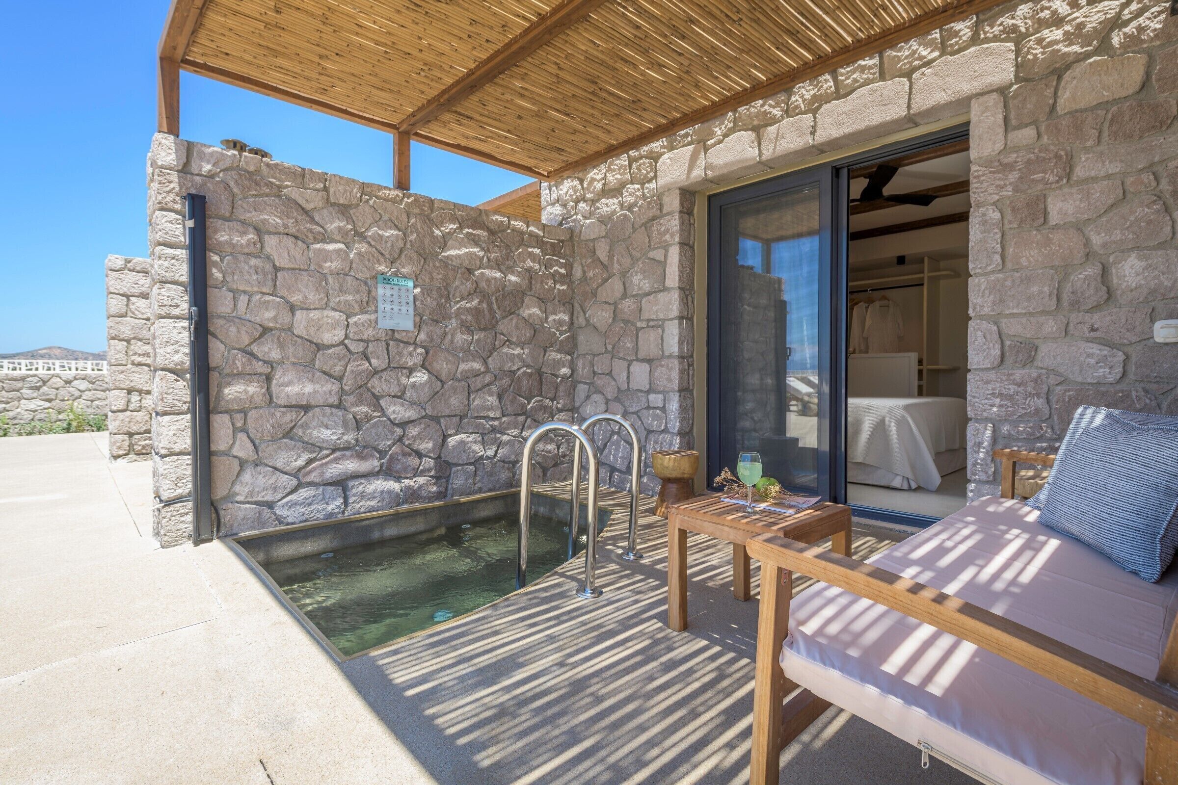 A stone-walled outdoor area with a pool, a wooden bench, and a glass door leading to a room.