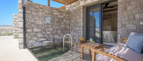 A stone-walled outdoor area with a pool, a wooden bench, and a glass door leading to a room.