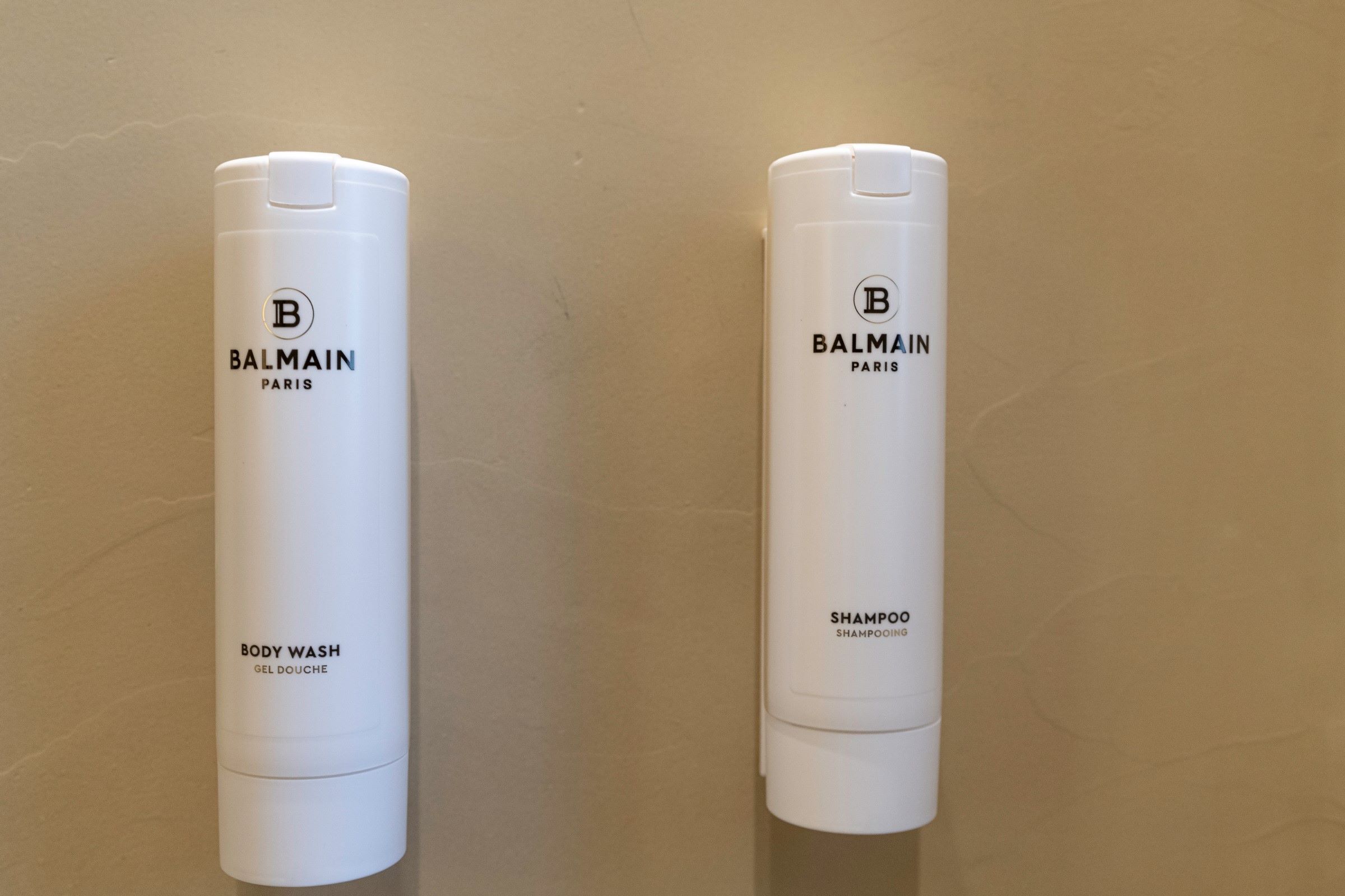 Two white Balmain Paris body wash and shampoo bottles against a plain wall.