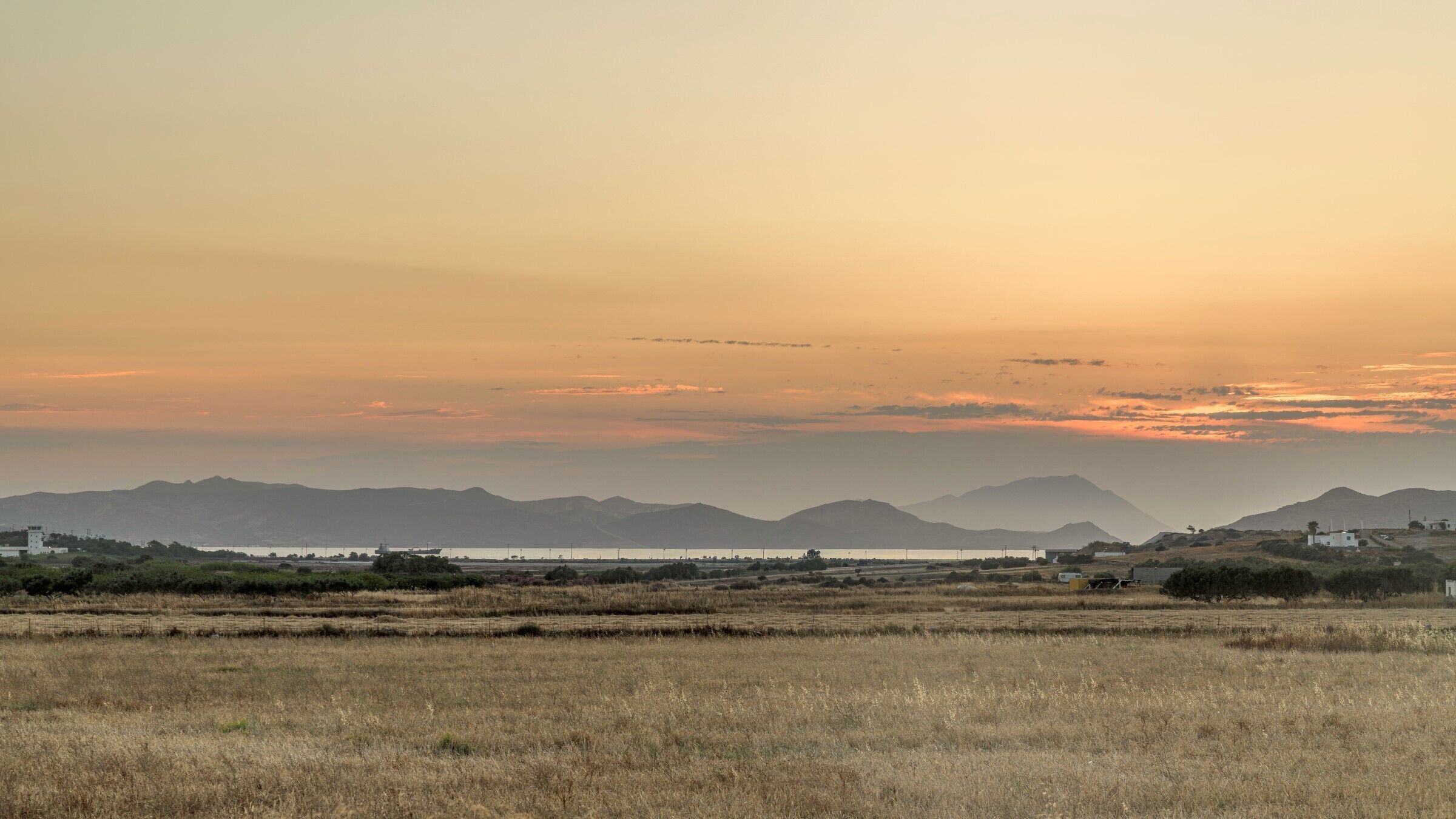 Un paisaje llano con campos de hierba seca, montañas a lo lejos y algunos edificios.