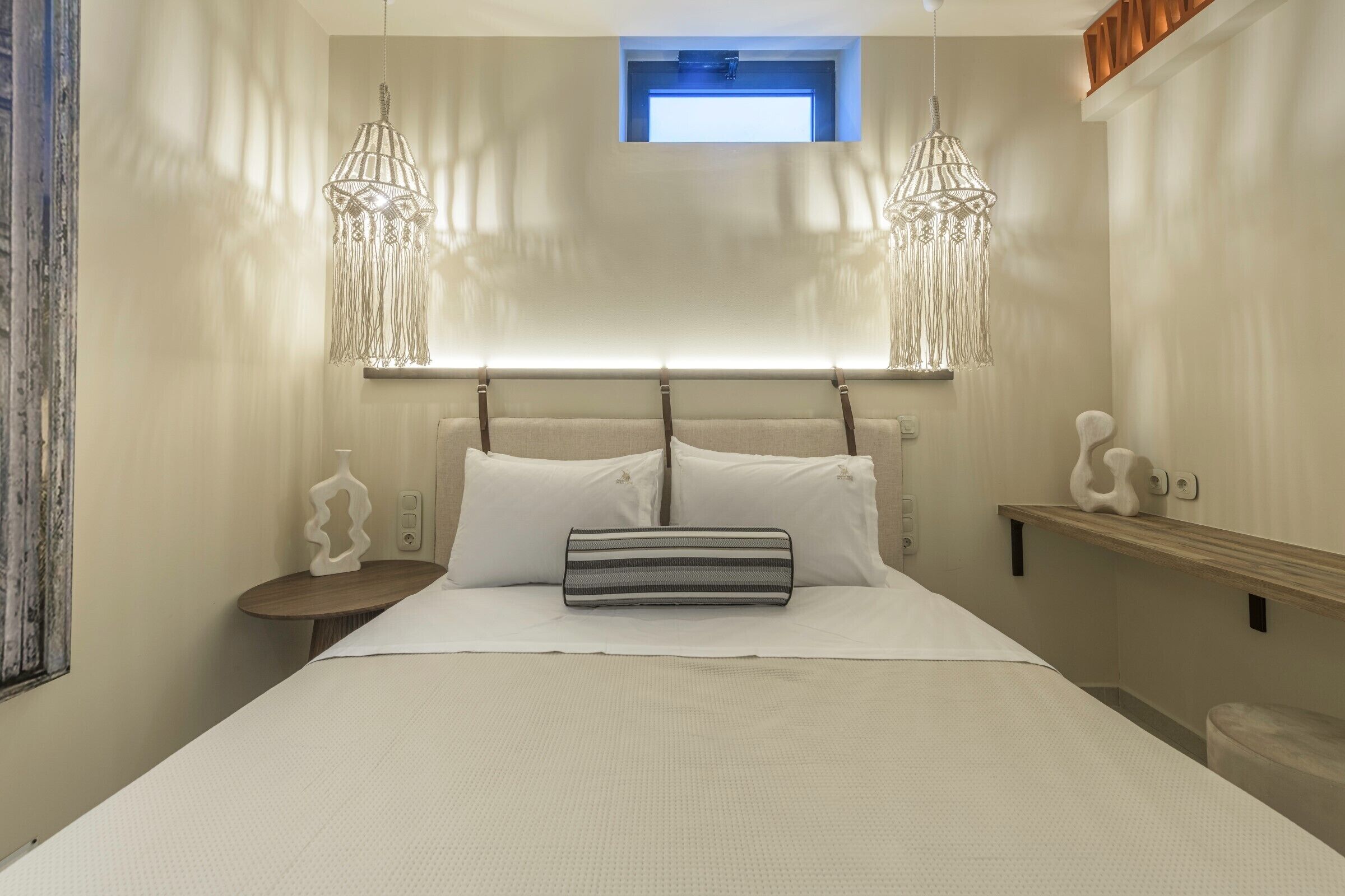 Uma cama bem arrumada com almofada listrada, duas luminárias pendentes, uma mesa de cabeceira com objeto decorativo e uma pequena janela sobre a cama.