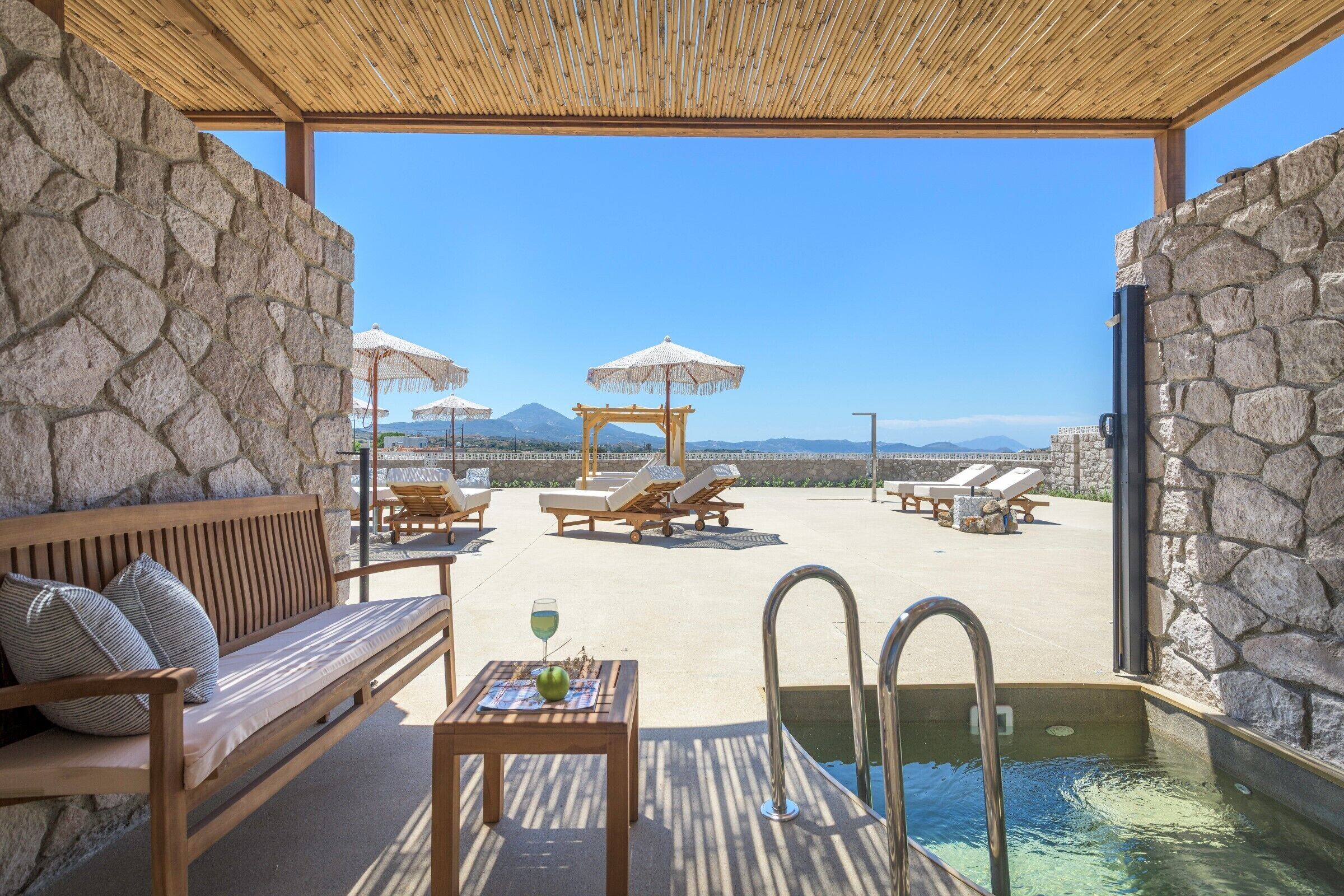 Zona de piscina con sillones reclinables, un banco de madera y una mesa con una bebida.