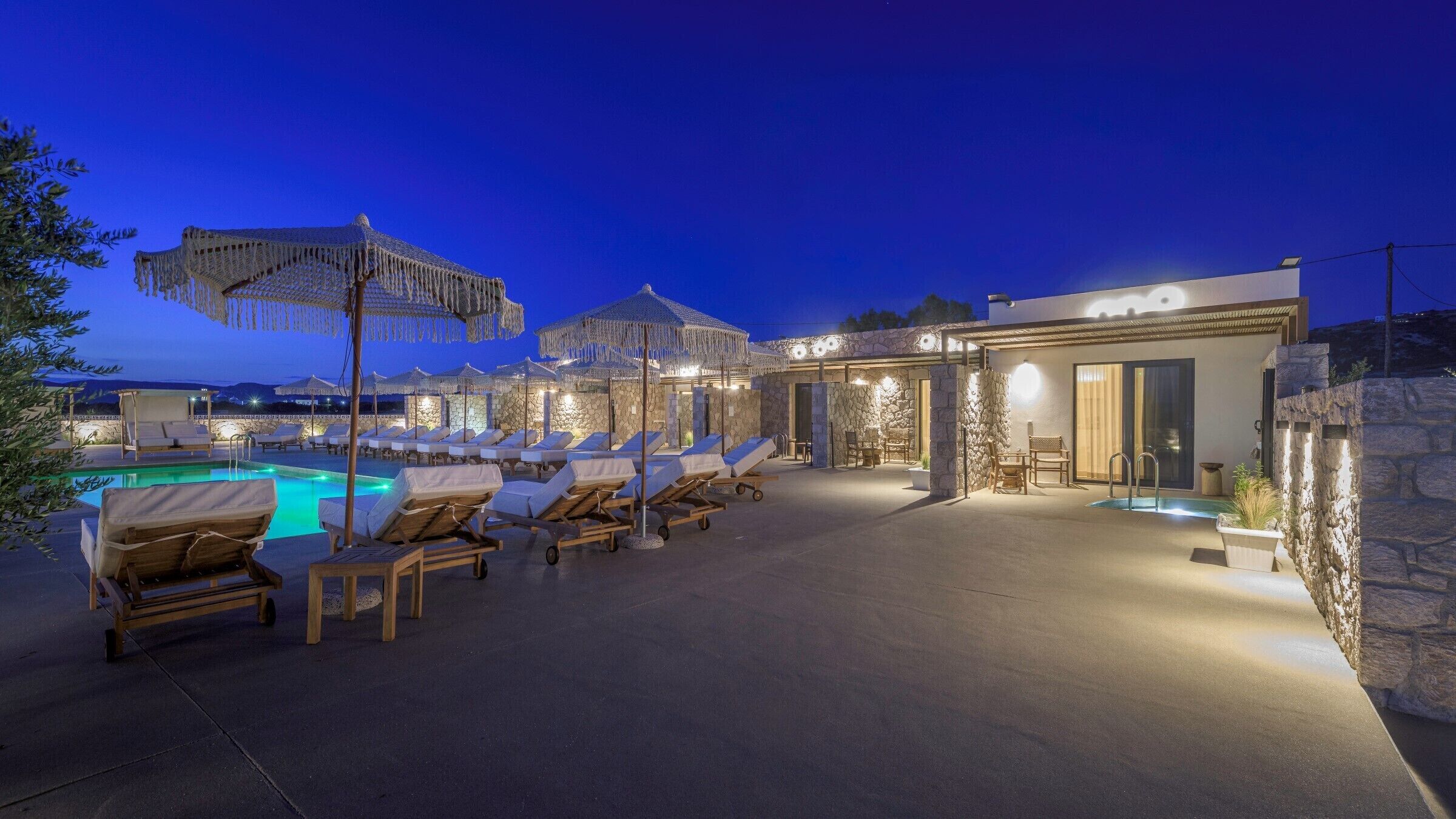 Un espace aménagé au bord de la piscine, avec des chaises longues et des parasols, un mur en pierre et un bâtiment moderne doté d’une porte en verre.