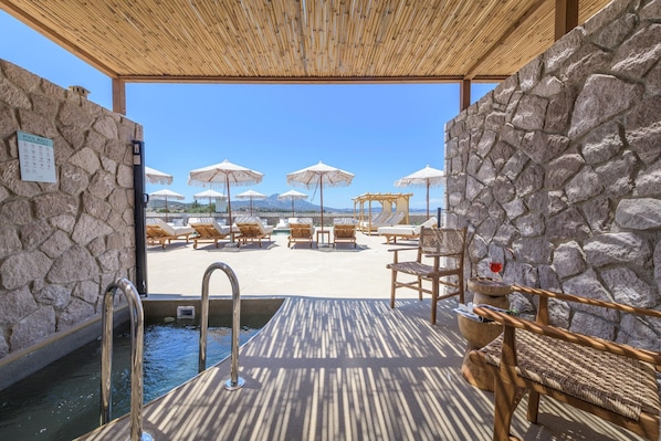 A pool area with stone walls, lounge chairs, umbrellas, and a wooden deck. - Zefyria Luxury Suites (Milos)